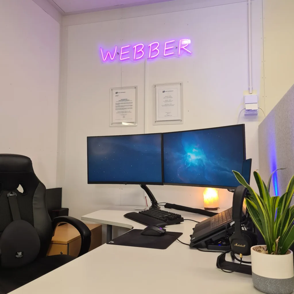 A professional home office workstation featuring dual monitors on an ergonomic monitor arm mount. The setup includes an ergonomic office chair, a low-profile keyboard, a warm salt lamp, and a snake plant against a white wall with framed certificates, showcasing a clean and organized workspace with minimal visible cables.