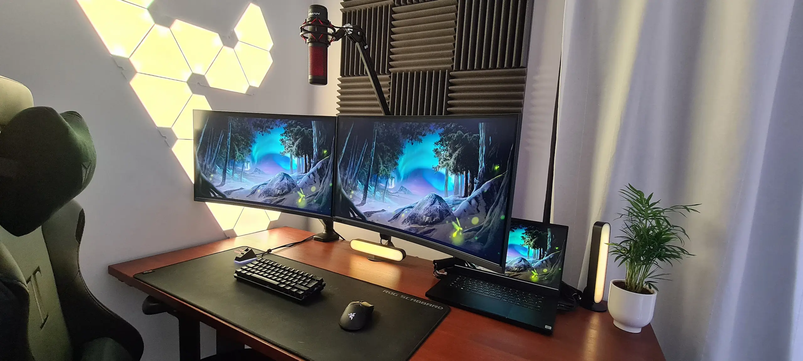 A clean and organized home office desk setup featuring dual monitors, a laptop, and a professional microphone on a boom arm. The workspace is enhanced by geometric LED wall panels, acoustic foam, and a small indoor plant, demonstrating a minimalist and ergonomic design with effective cable management.