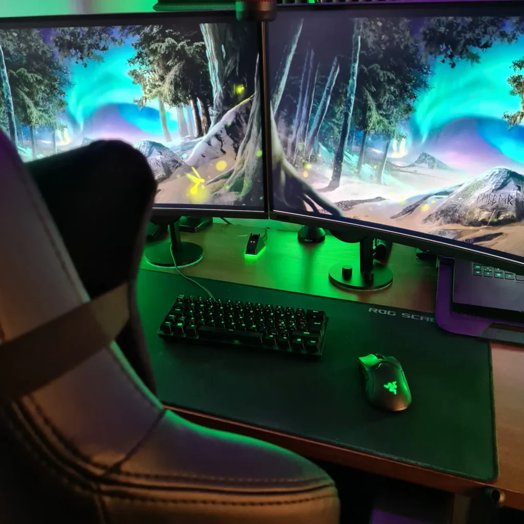 An immersive point-of-view shot of a clean gaming workstation featuring dual monitors, a black mechanical keyboard, and a gaming mouse on a large desk mat. The workspace is illuminated by vibrant green under-desk RGB lighting, highlighting an organized and cable-managed setup.