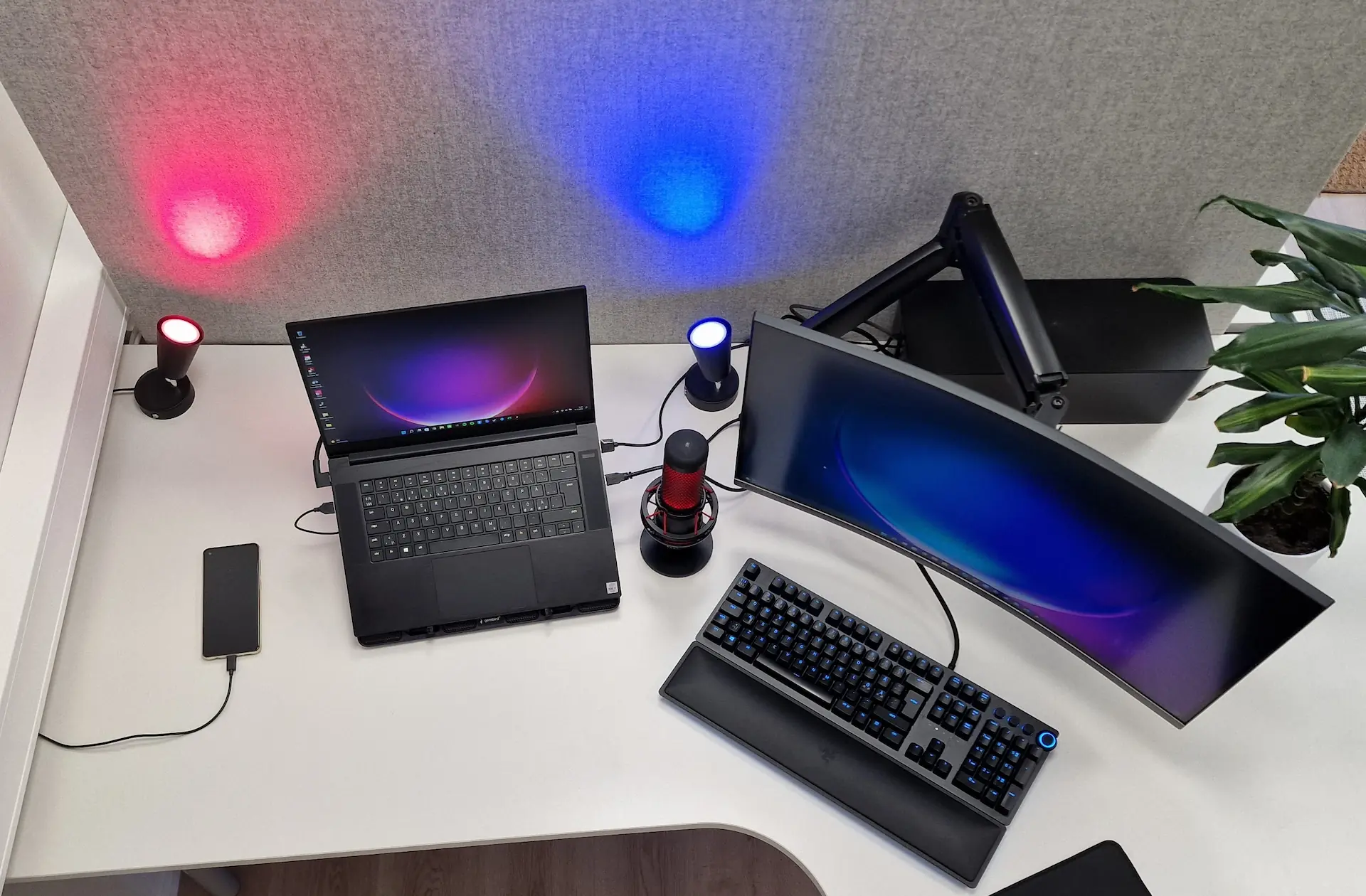 Top-down view of a white home office desk featuring a curved ultrawide monitor on a mounting arm, an open laptop, and a mechanical keyboard. A black cable management box sits in the corner to hide cords, complemented by vibrant red and blue LED accent lighting that creates a high-performance gaming or work atmosphere.